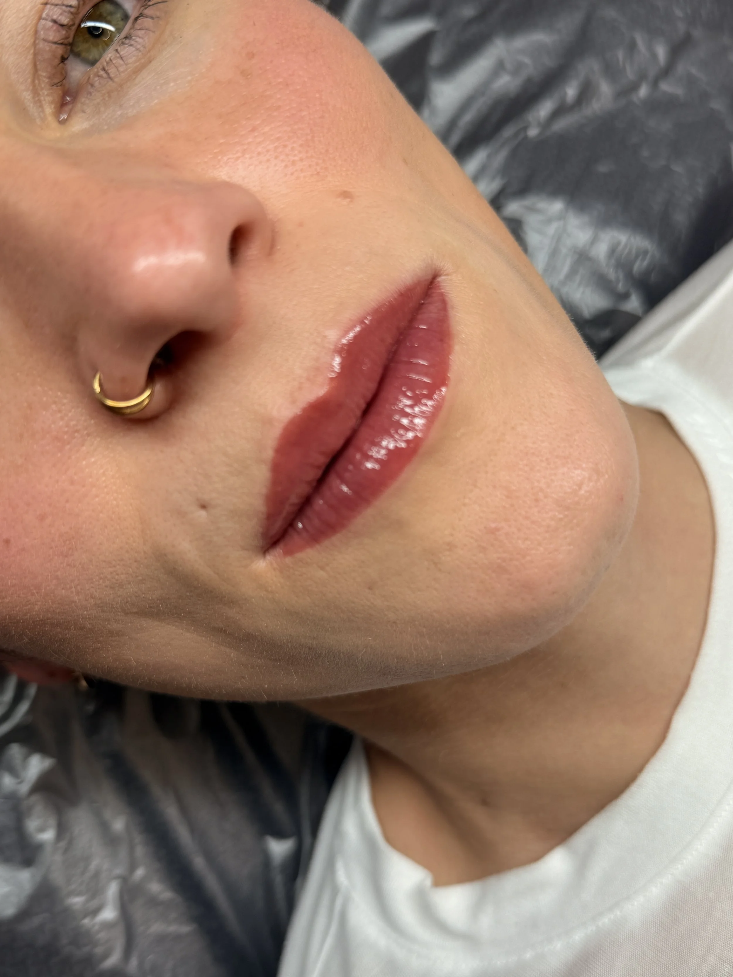 Nahaufnahme von einem Gesicht mit Fokus auf Lippen, Nase und einem Auge, mit goldener Nase-Niete, im Hintergrund dunkel und grau. Ein Teil eines weißen Shirts ist sichtbar.