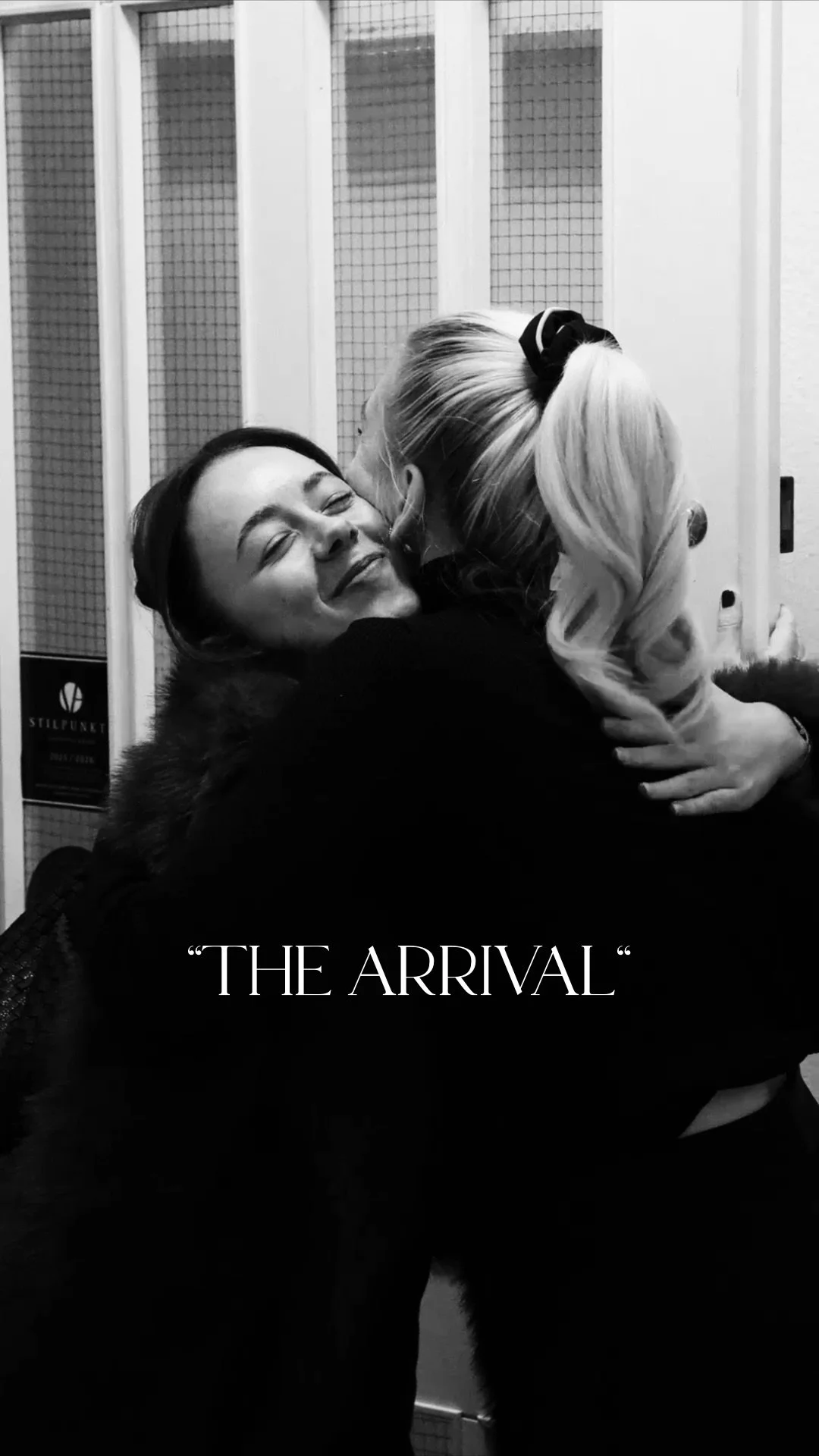 Zwei Frauen umarmen sich herzlich und lachen in einem öffentlichen Raum. Der Text auf dem Bild lautet "The Arrival".