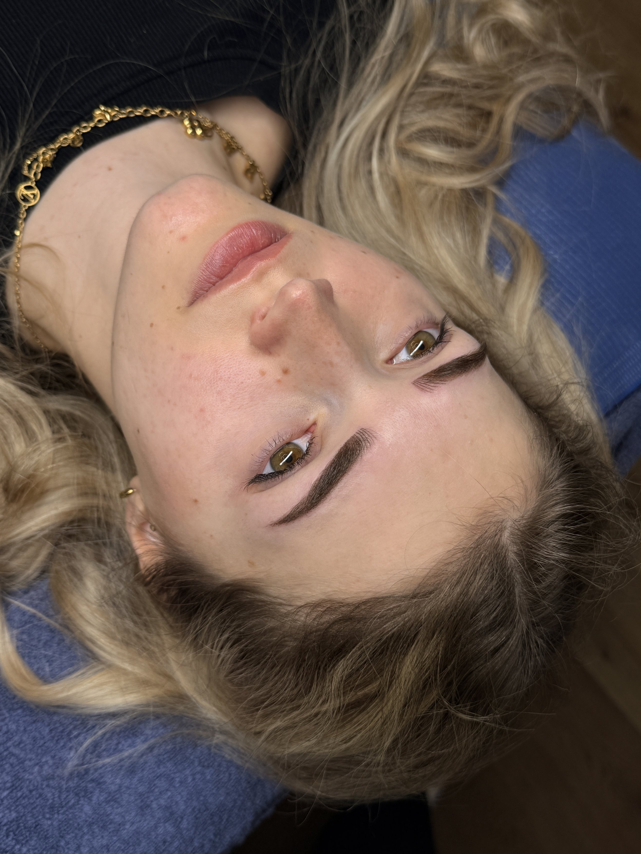 Nahaufnahme eines jungen Mädchens mit blonden, lockigen Haaren, braunen Augen, Sommersprossen auf den Wangen und Lippen mit natürlichem Make-up, trägt eine goldene Halskette, liegt auf dem Rücken auf einer blauen Oberfläche.