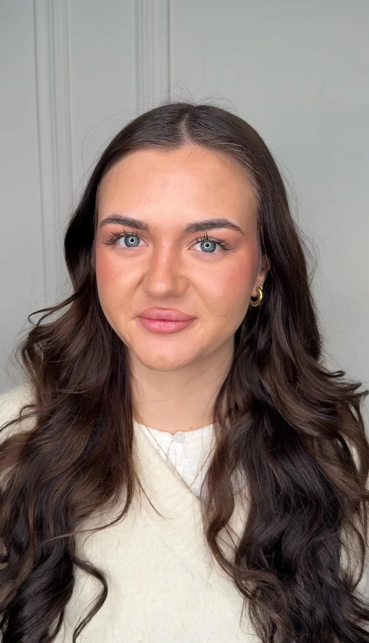 Nahaufnahme eines jungen Mädchens mit braunen, langen, lockigen Haaren, blauen Augen, leichtem Make-up, einer goldenen Ohrring und einem cremefarbenen Pullover vor einem grauen Hintergrund.