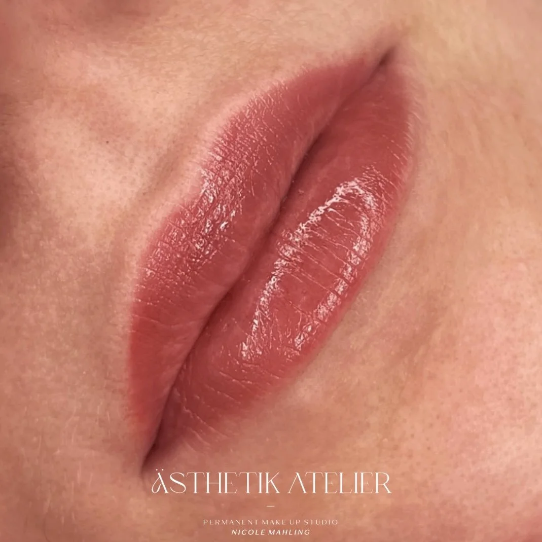 Nahaufnahme von vollen, gepflegten Lippen mit glossigem Make-up. Der Hintergrund ist unscharf, das Bild trägt den Text 'ÄSTHETIK ATELIER' und 'PERMANENT MAKE UP STUDIO NICOLE MAHLING'.