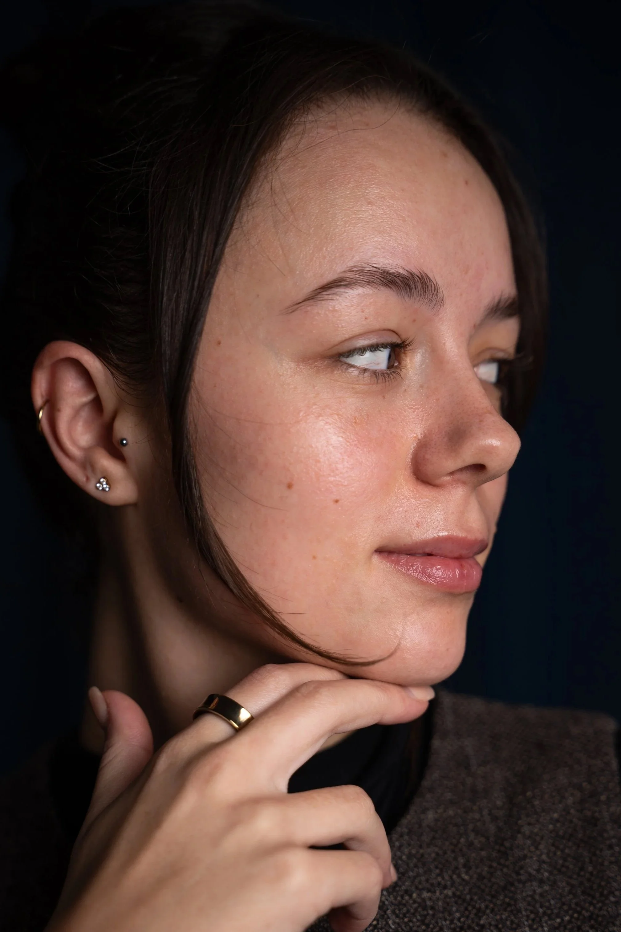 Nahaufnahme eines Frauengesichts mit glatter Haut, dunklen Haaren und mehreren Ohrpiercings, die die rechte Seite des Gesichts zeigt, mit einem dunklen Hintergrund.