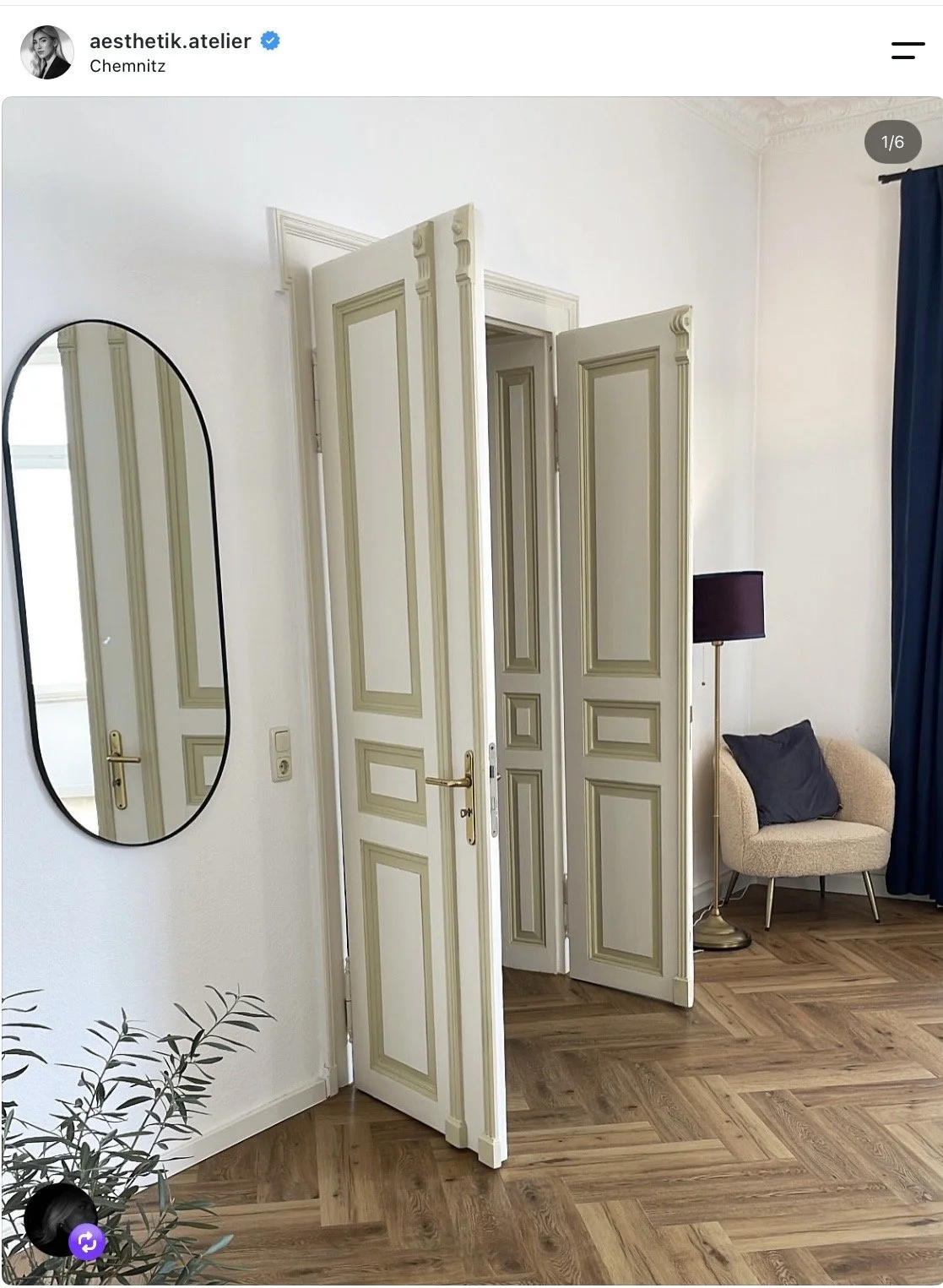 Ein Raum mit weißen Wänden und einer Holzfußboden. Im Mittelpunkt steht ein offener, dekorativ gestalteter Raumteiler mit weißen Türen und grünen Akzentlinien. Auf der linken Seite ist ein ovaler Spiegel an der Wand. Auf der rechten Seite befindet sich eine beige Sessel mit einem schwarzen Kissen, neben einem schwarzen Lampenschirm auf einem Goldständer. Einen dunklen blauen Vorhang sieht man an der rechten Seite.
