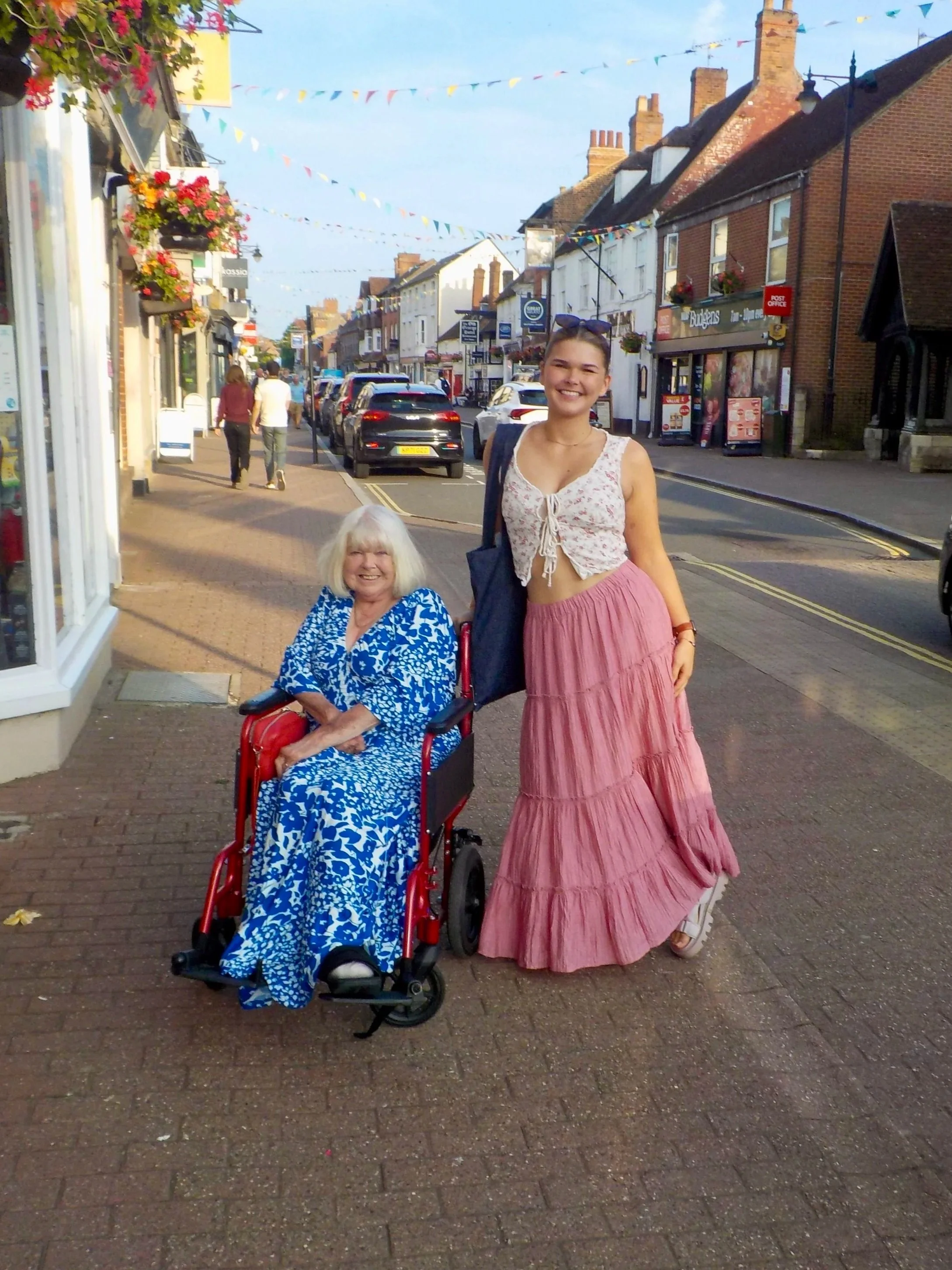 Anna and her mum. Anna is standing in a pink dress and a summer top. She has brown hair. On the left, her mum sits in her red wheelchair in a blue and white patterned dress. She has blonde hair.