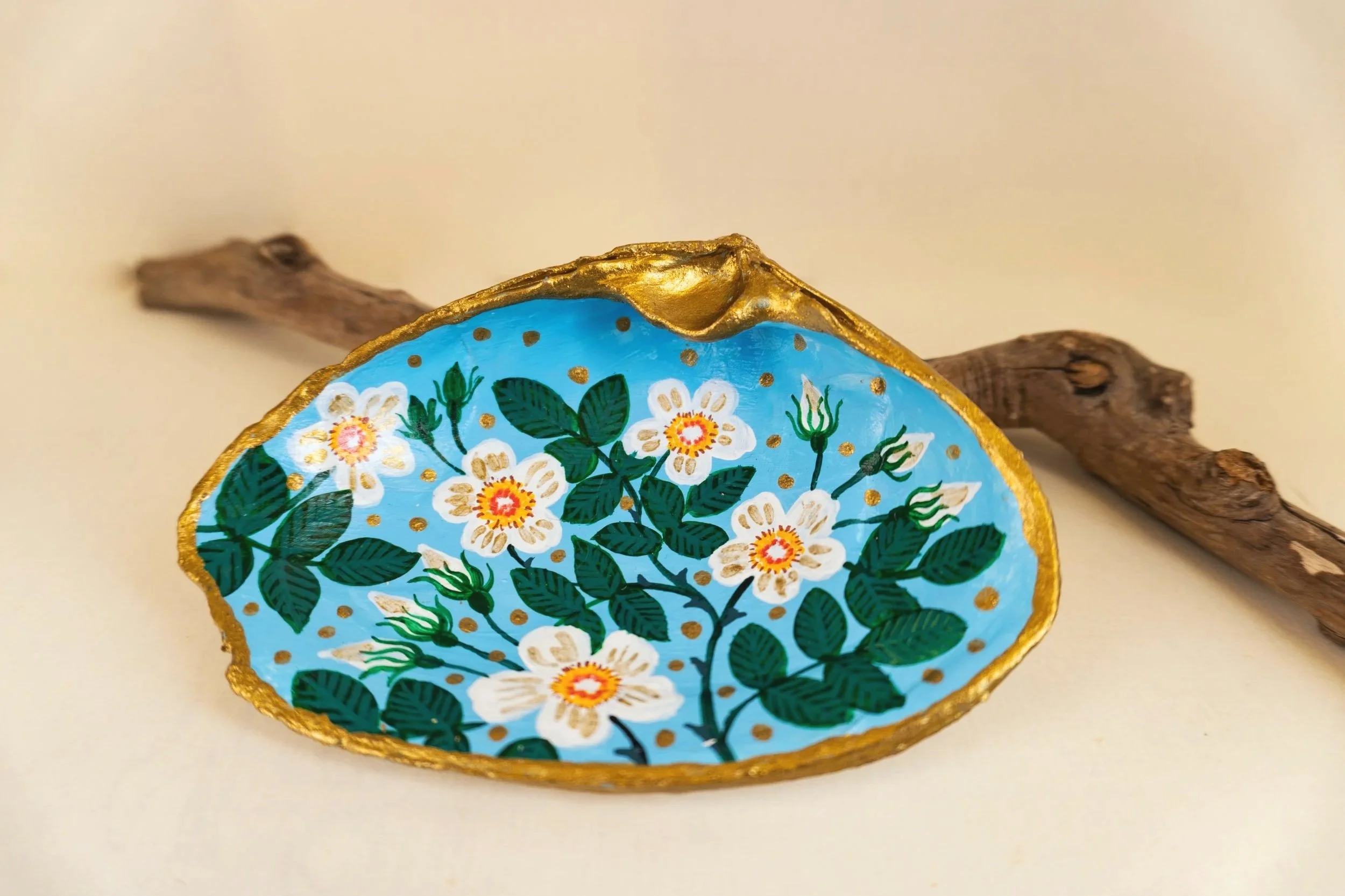 Decorative oval object painted with white flowers and green leaves on a light blue background, outlined with gold, and placed on a piece of wood.