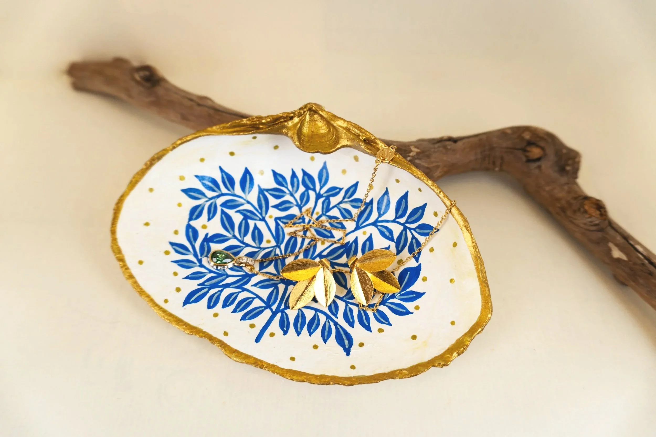 A decorative jewelry dish with a blue leaf pattern, gold trim, and gold jewelry including a necklace with leaf-shaped pendant resting inside.