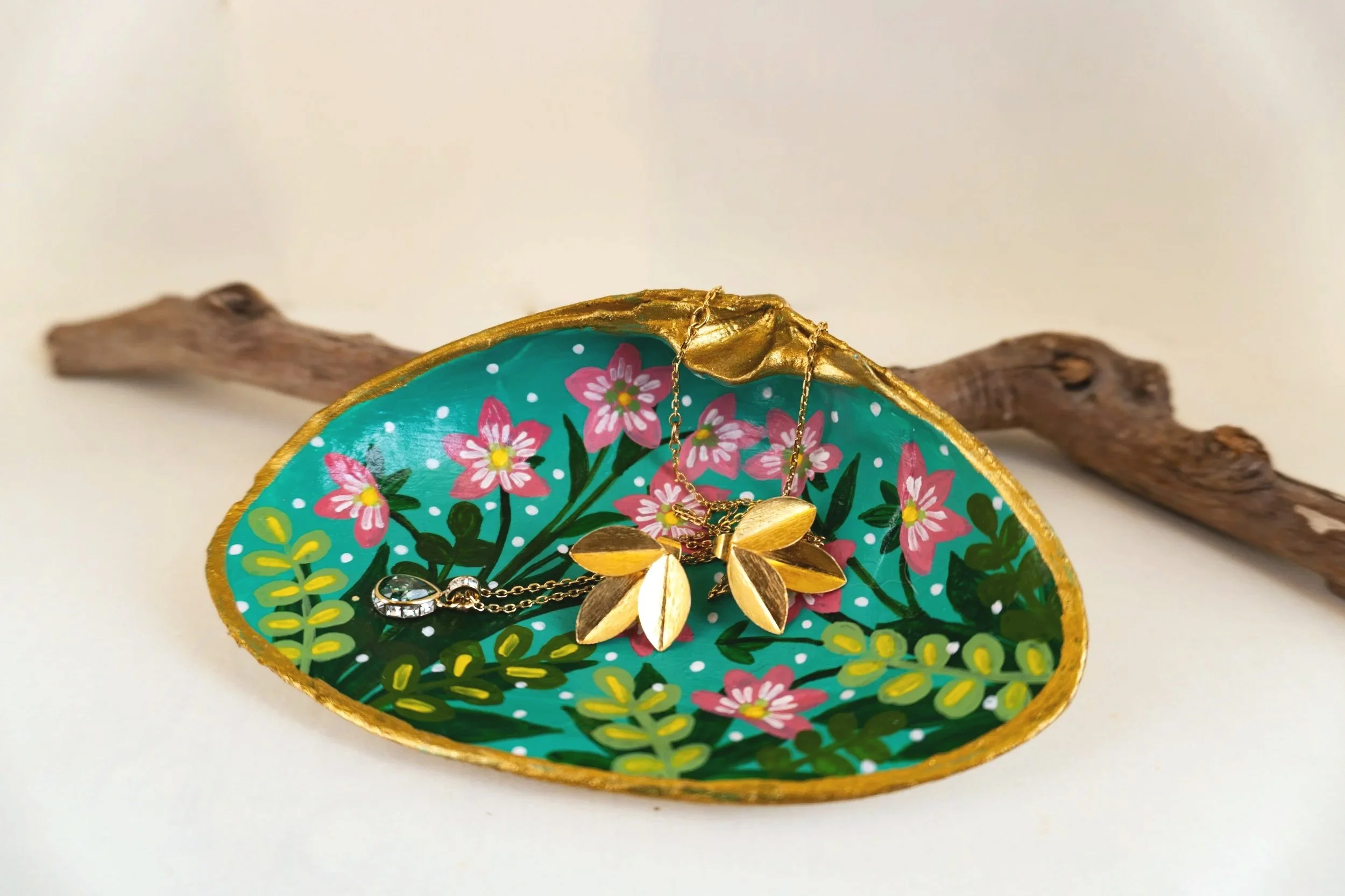 A painted decorative shell with pink, yellow, and green floral patterns, gold trim, and jewelry including gold leaf-shaped earrings and layered necklaces displayed inside.
