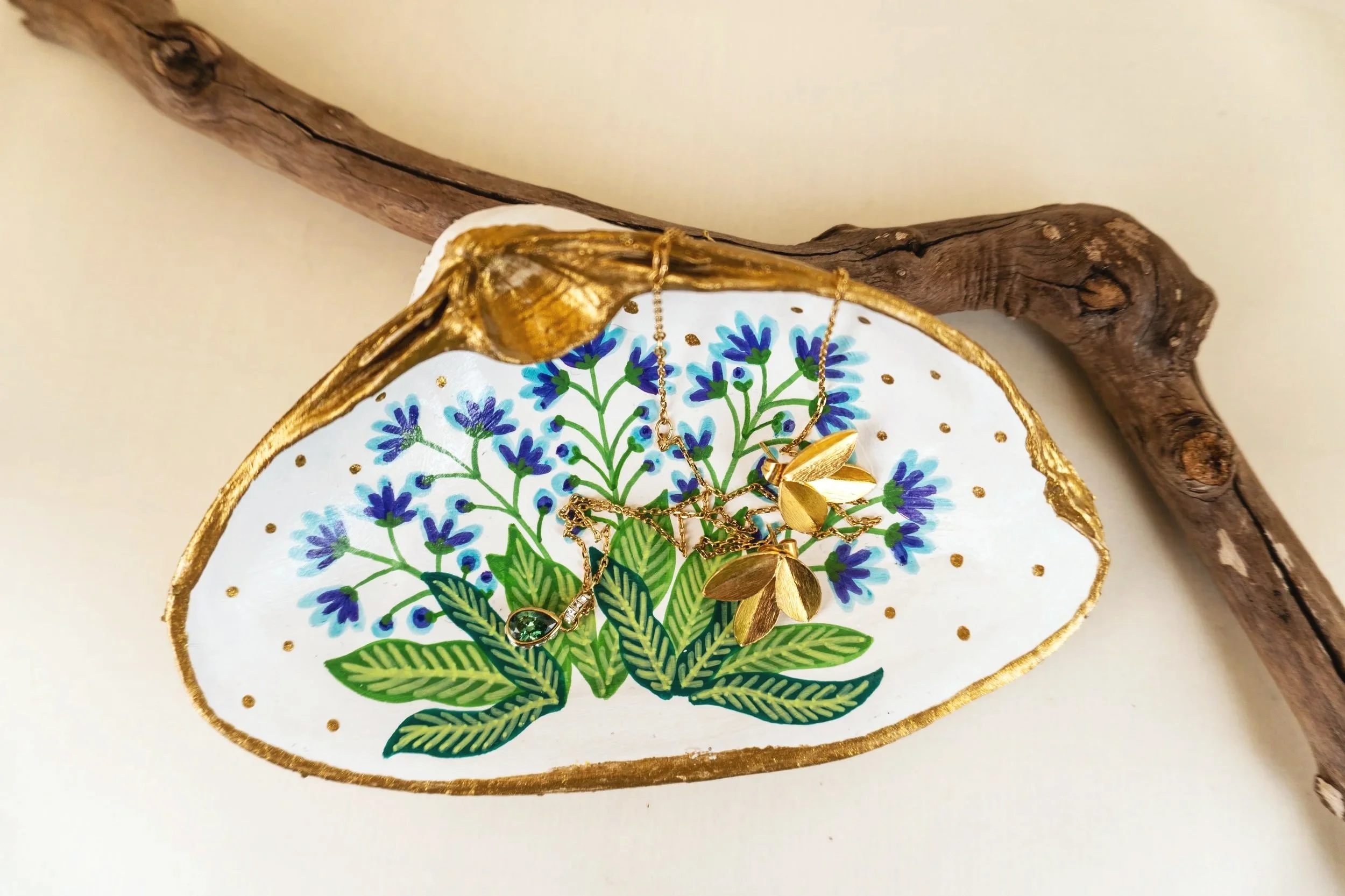Gold jewelry, including necklaces and earrings, laid on a decorative plate with a floral design, placed on a white surface with a piece of twisted wood in the background.