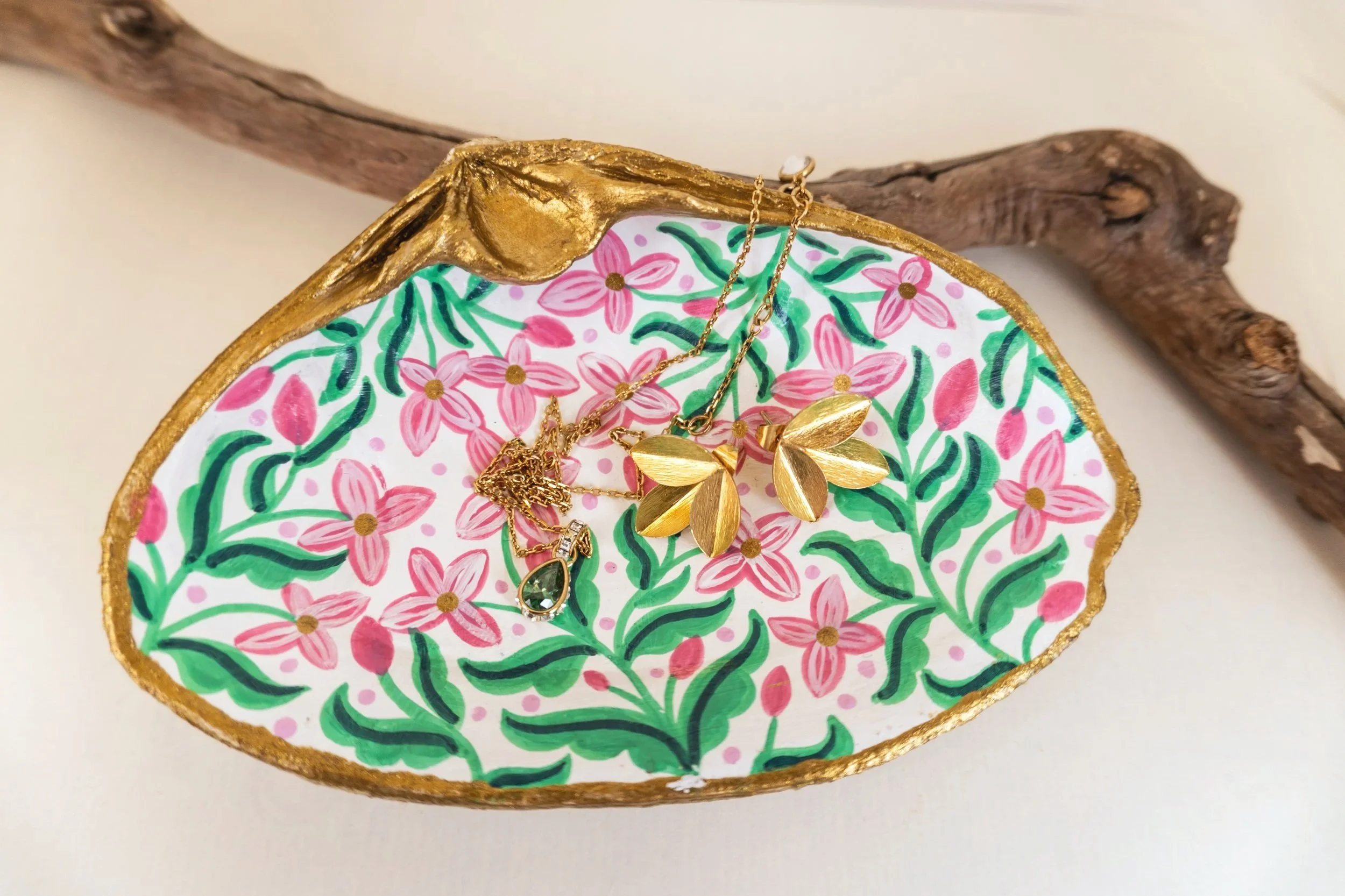 A decorative oval-shaped tray with pink floral and green leaf pattern, trimmed with gold, holding gold-colored jewelry including necklaces with leaf pendants and a green gemstone charm, placed on a white surface with a wooden branch in the background.