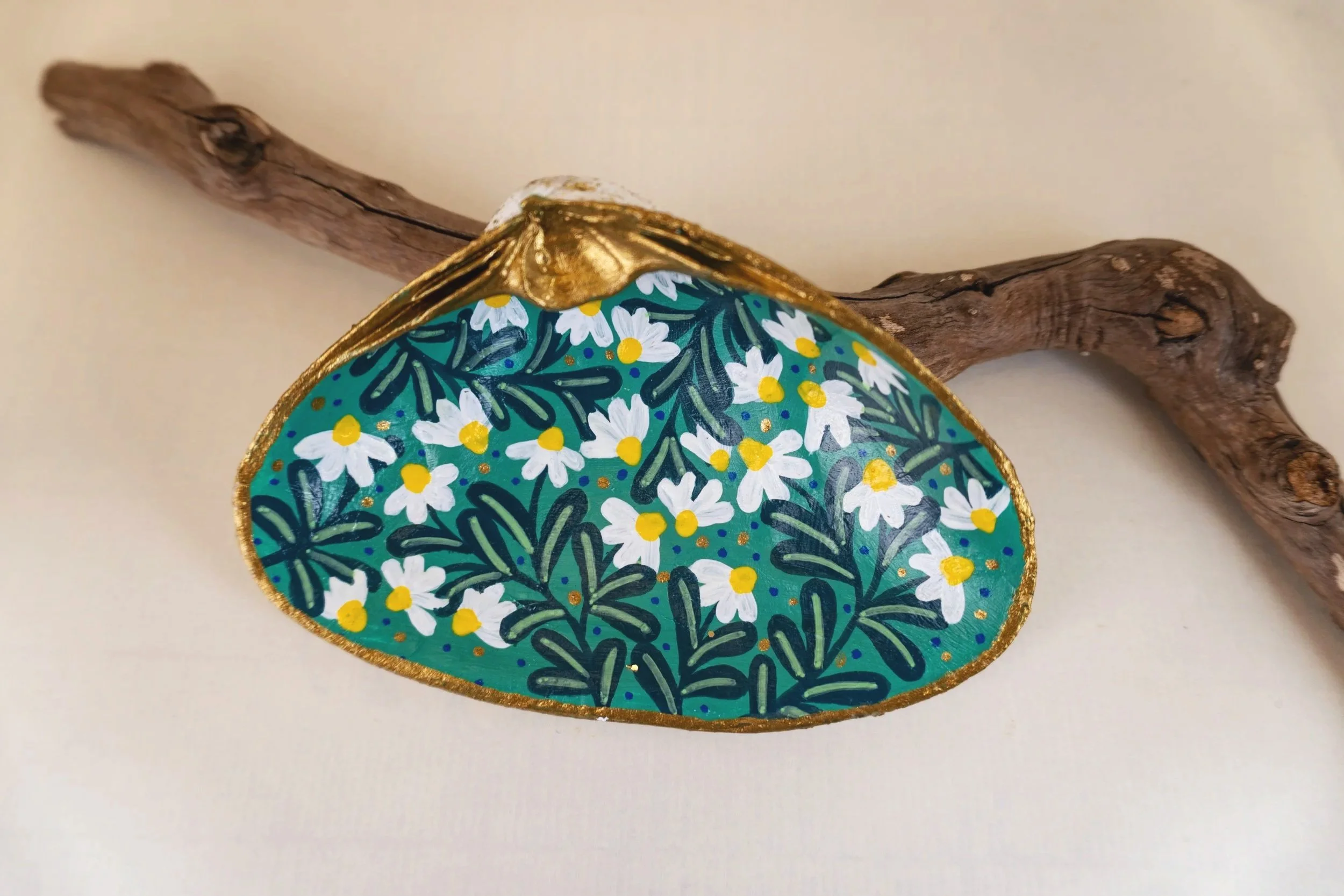 Decorative oval object with a floral pattern of white flowers with yellow centers and dark green leaves, edged with gold, resting on a piece of driftwood.