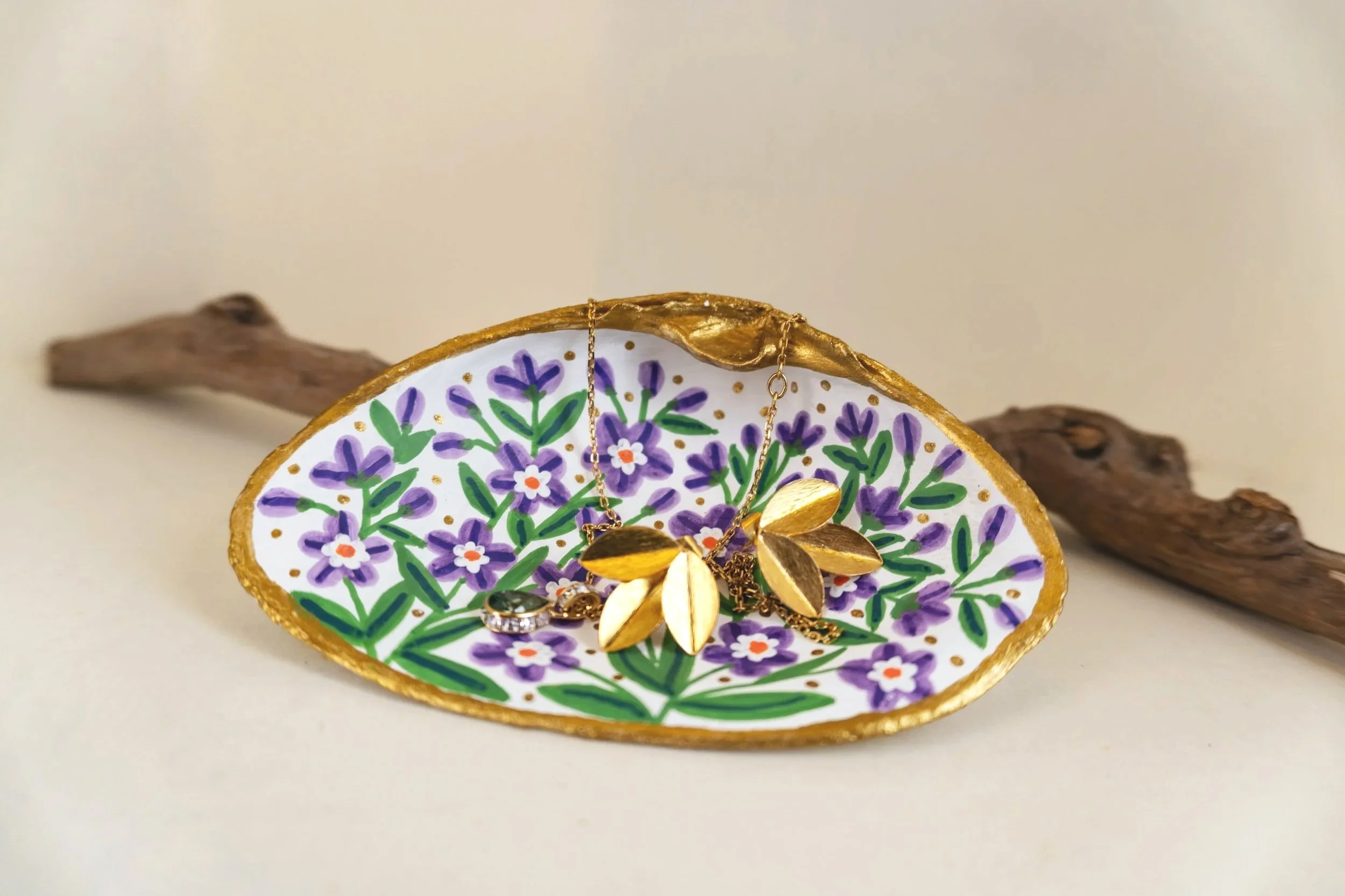 Decorative ceramic dish with purple flowers and green leaves design, gold trim, holding gold jewelry including a leaf-shaped pendant and rings, on a white surface with driftwood in the background.