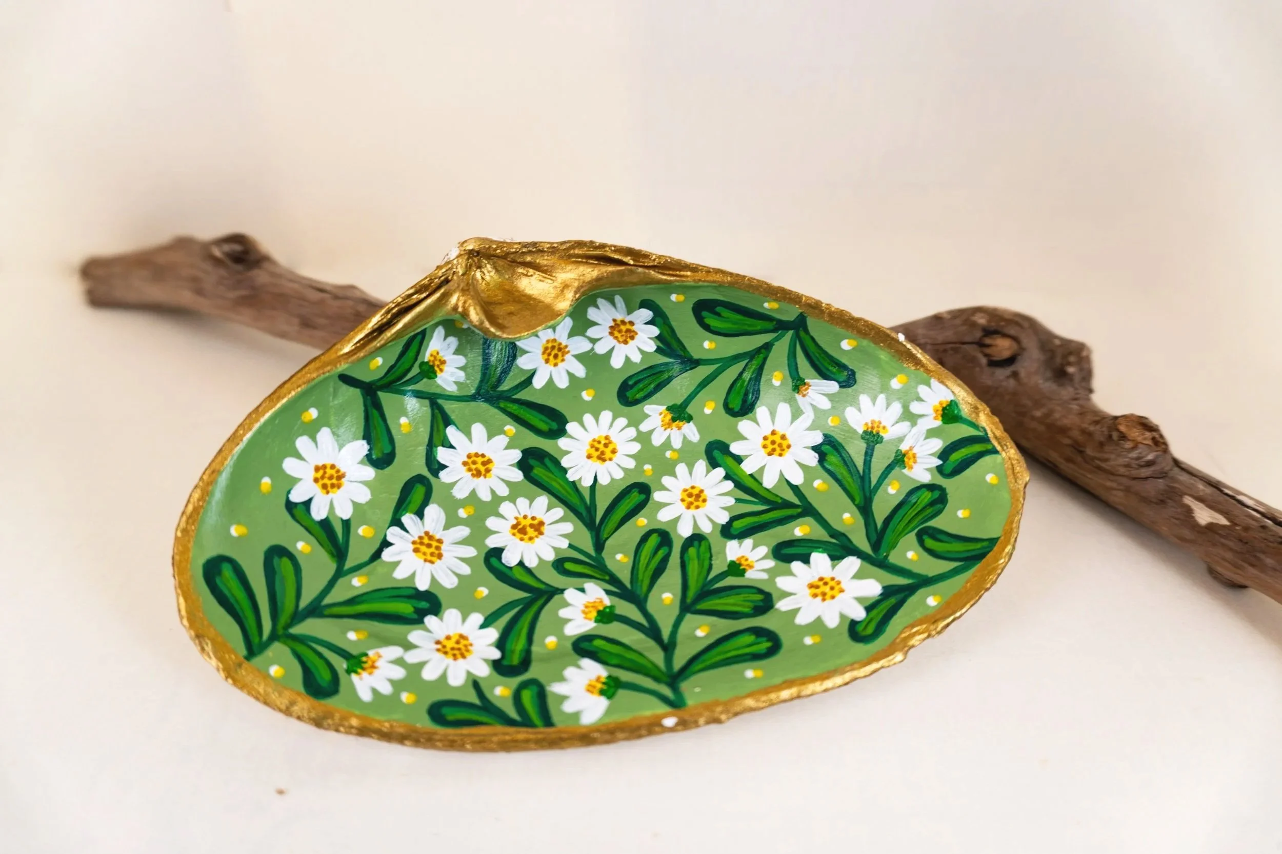 A decorative painted stone with a floral pattern of white flowers and green leaves, outlined in gold, resting on a wooden stick.
