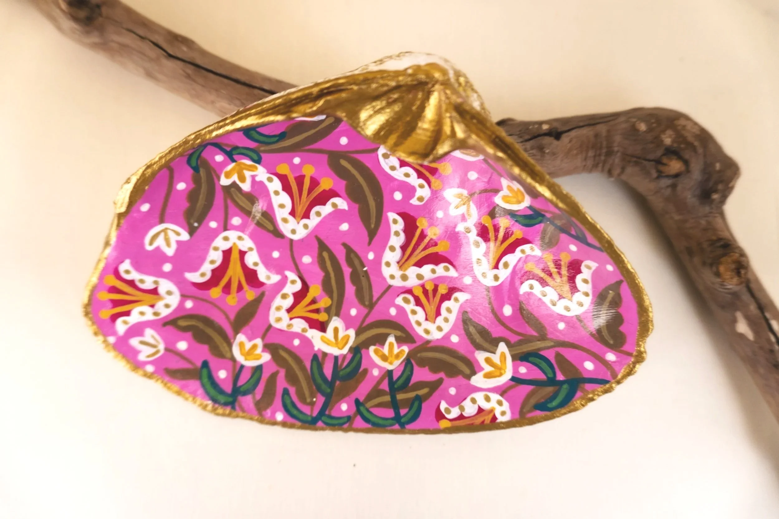 Close-up of a decorative, painted rock with a vibrant pink background and intricate floral patterns in white, gold, green, and brown, resting against a piece of driftwood.