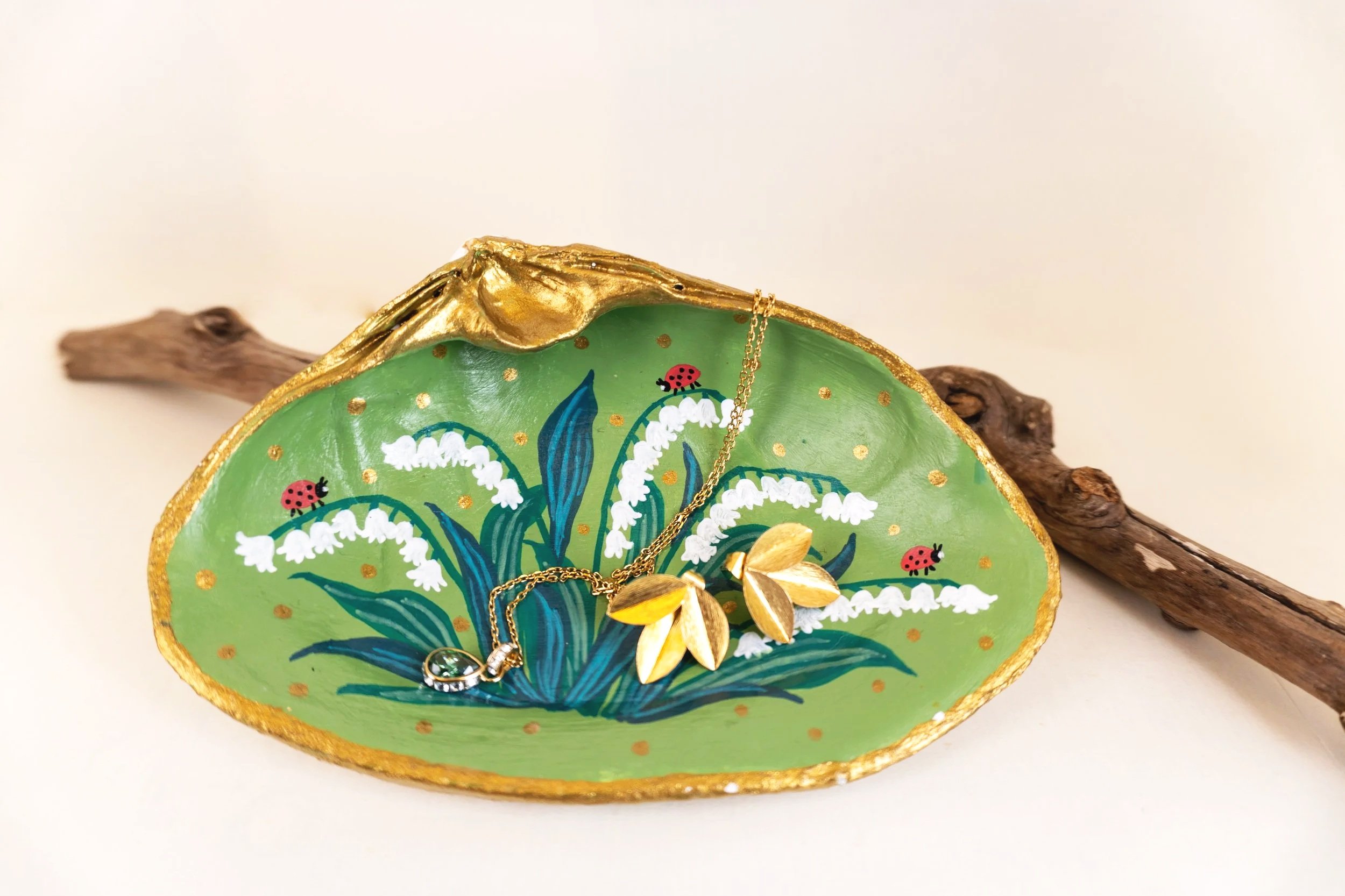 Decorative ceramic dish with green base color, painted with white flowers, blue leaves, and small red ladybugs, with gold trim on the edges, placed on a piece of driftwood.