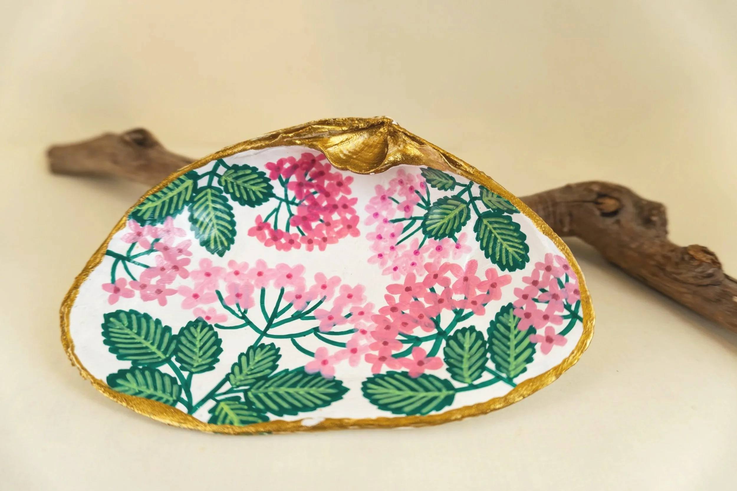 Decorative plate with pink floral pattern and green leaves, gold trim, resting on a piece of driftwood on a cream background.