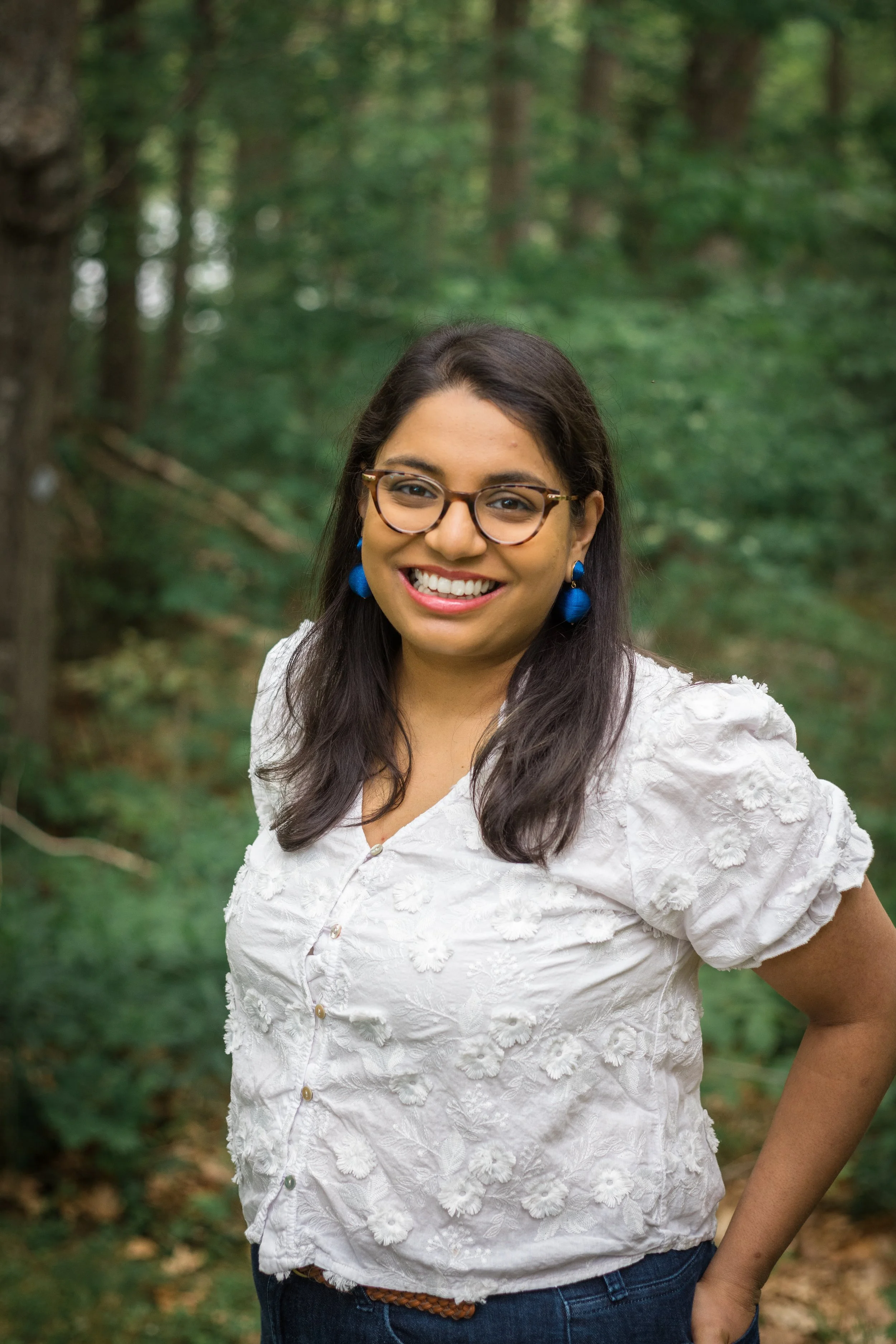 A woman with dark hair, glasses, and a white floral blouse smiling outdoors in a forested area.