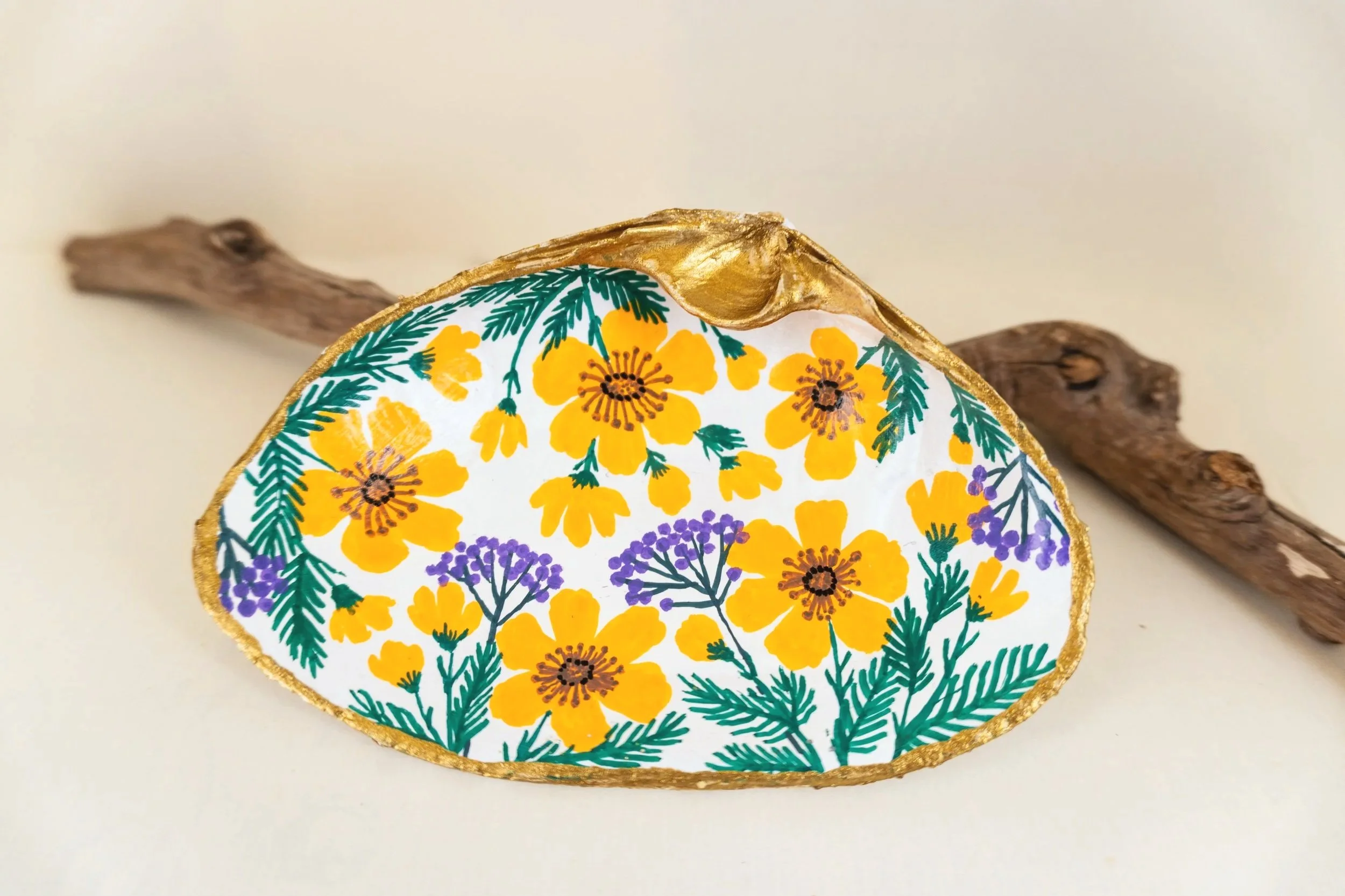 Decorative oval object with a floral pattern featuring yellow, purple, and green colors, edged with a gold trim, resting on a wooden branch against a plain background.