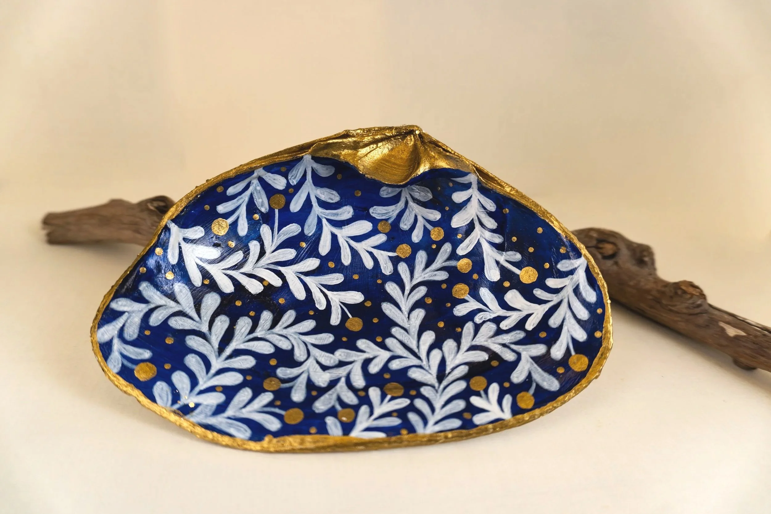 A decorative wooden piece painted with a blue background and white leaf pattern, edged with gold, and resting on a piece of driftwood.