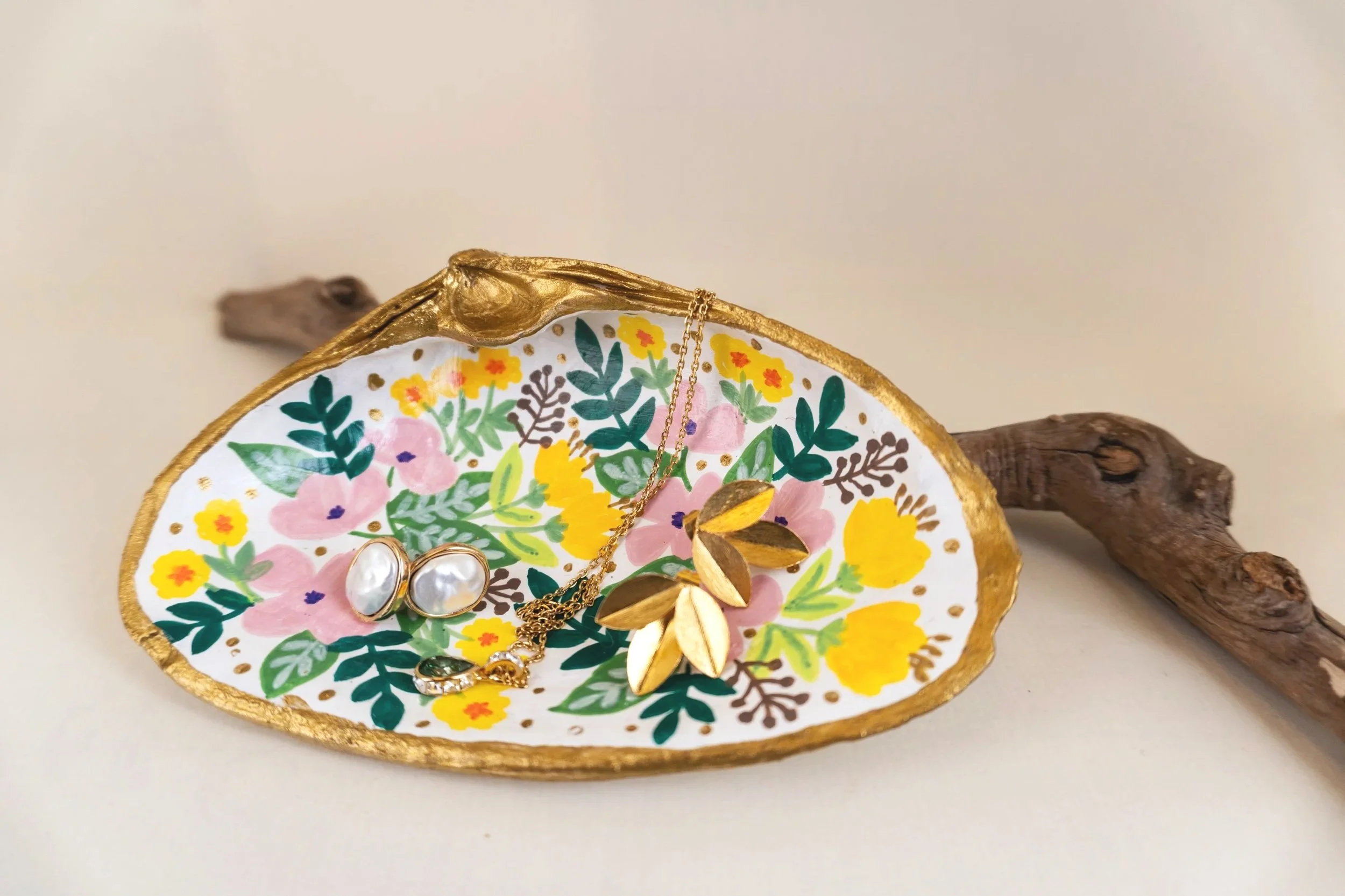 Decorative ceramic dish with colorful floral pattern, holding gold jewelry including a leaf-shaped necklace, a pearl ring, and a chain. The dish is placed on a white surface with wood pieces as background.