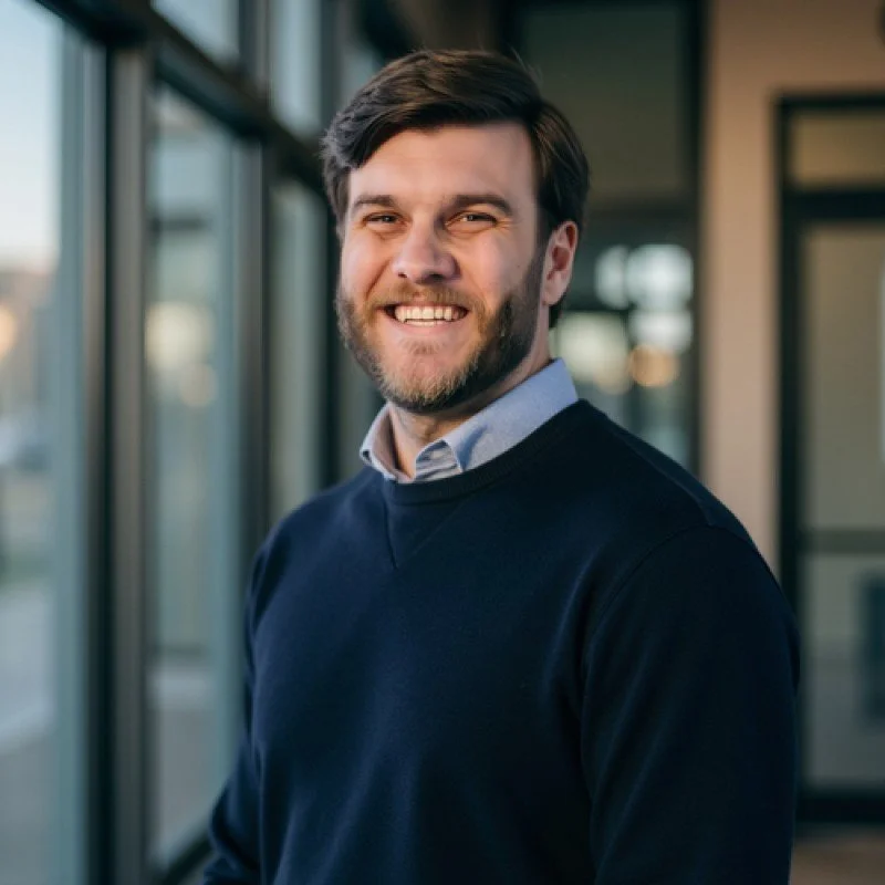Geoff Mader, a smiling man with brown hair and a beard, wearing a light blue collared shirt and a navy sweater, standing indoors near large windows with a modern building in the background.