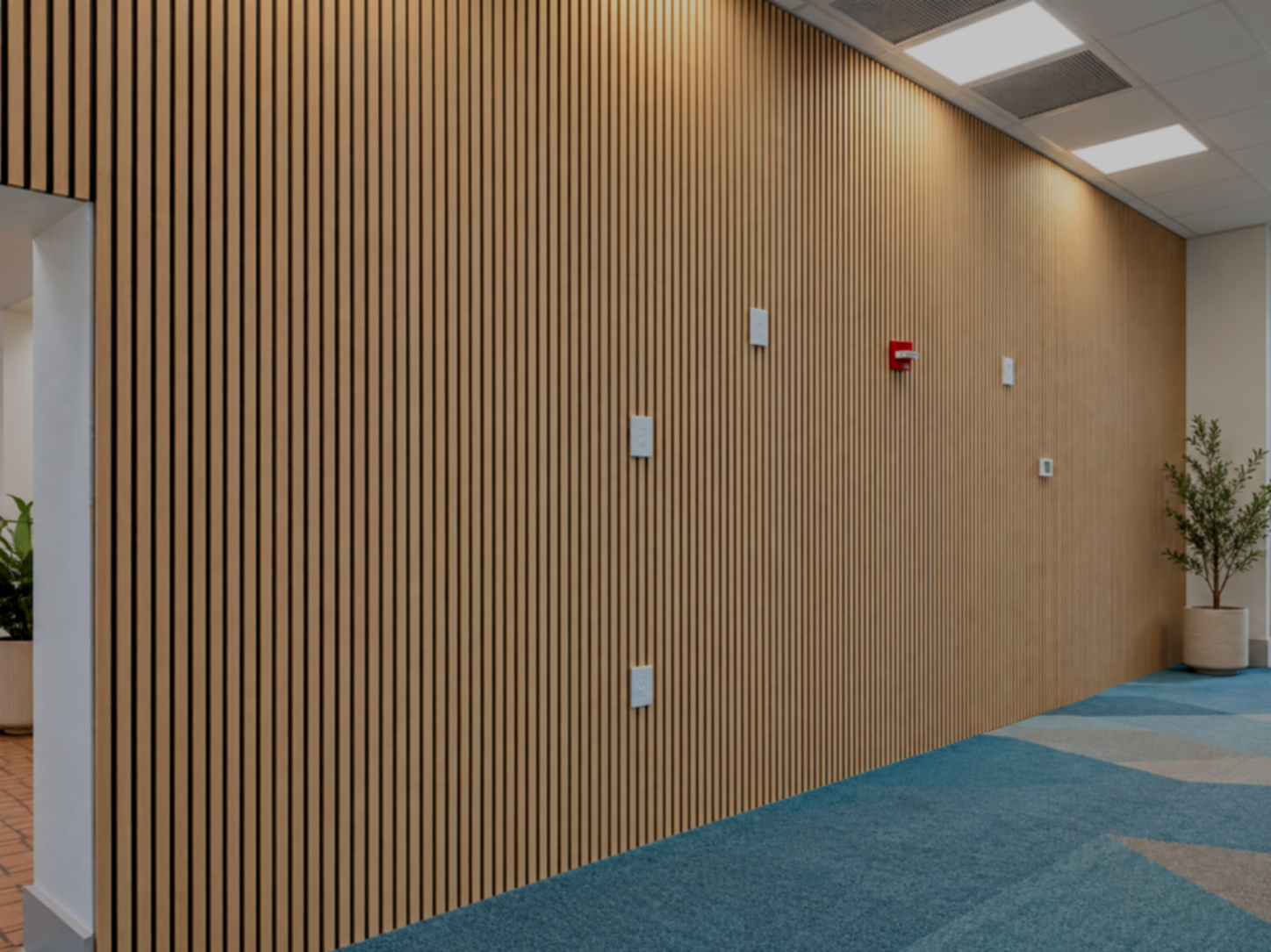 Interior office wall with vertical wooden slats, white and red fire alarm, thermostat, potted plant, and modern ceiling lighting.