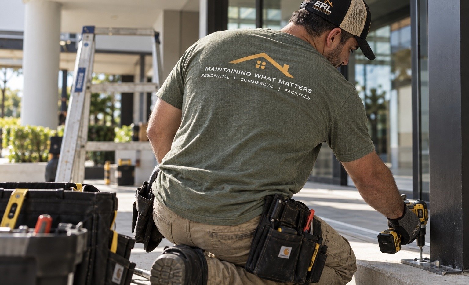 A worker installing a metal post on a concrete surface outside a building, using a cordless drill. He is wearing a green T-shirt with a logo and text about maintenance services, cargo pants with a tool belt, and a cap. There are tools and equipment nearby, including a tool bag and a ladder in the background.