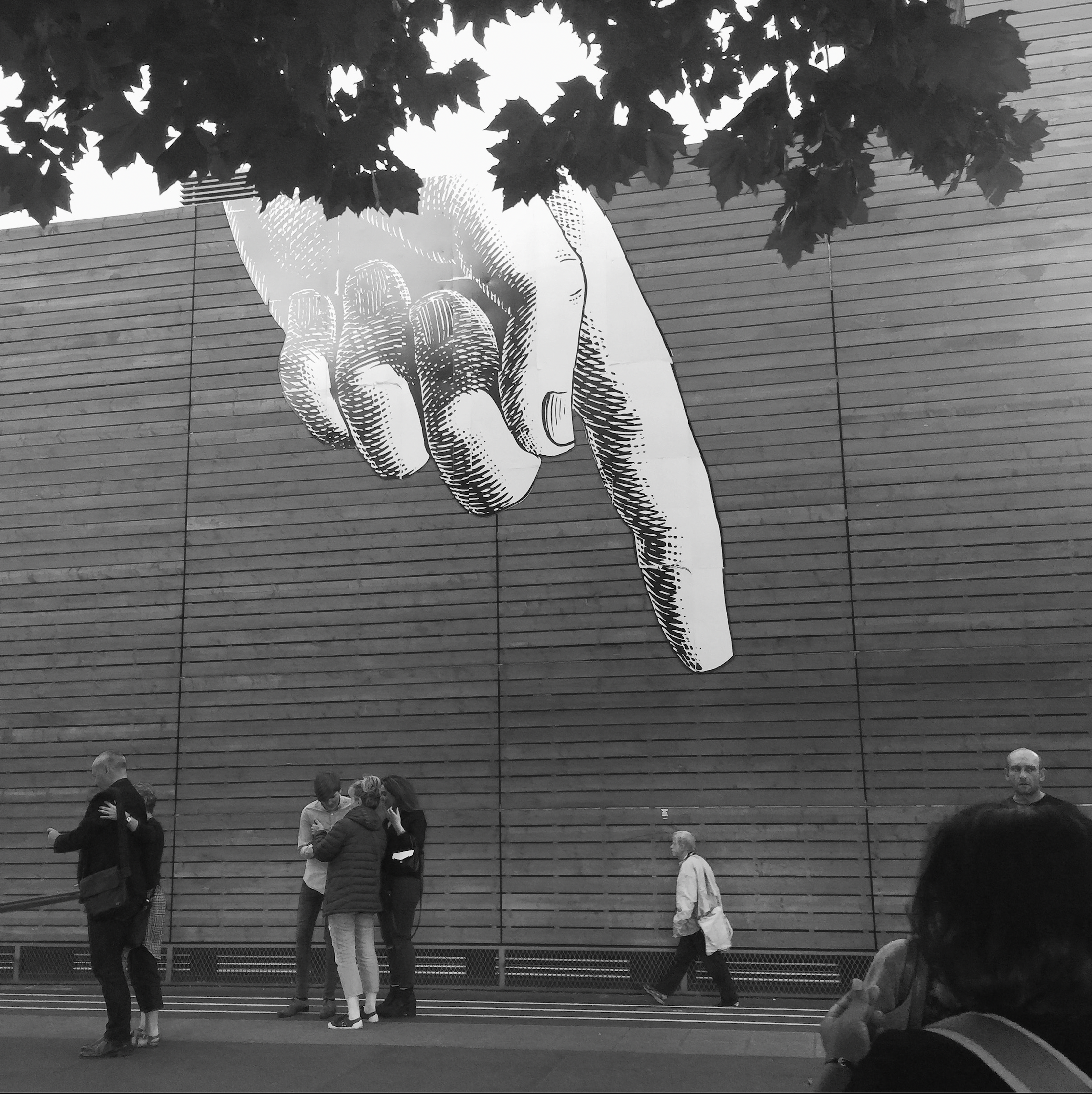 A black and white photograph of a large mural of a hand with a finger pointing down as a pedestrian walks underneth.