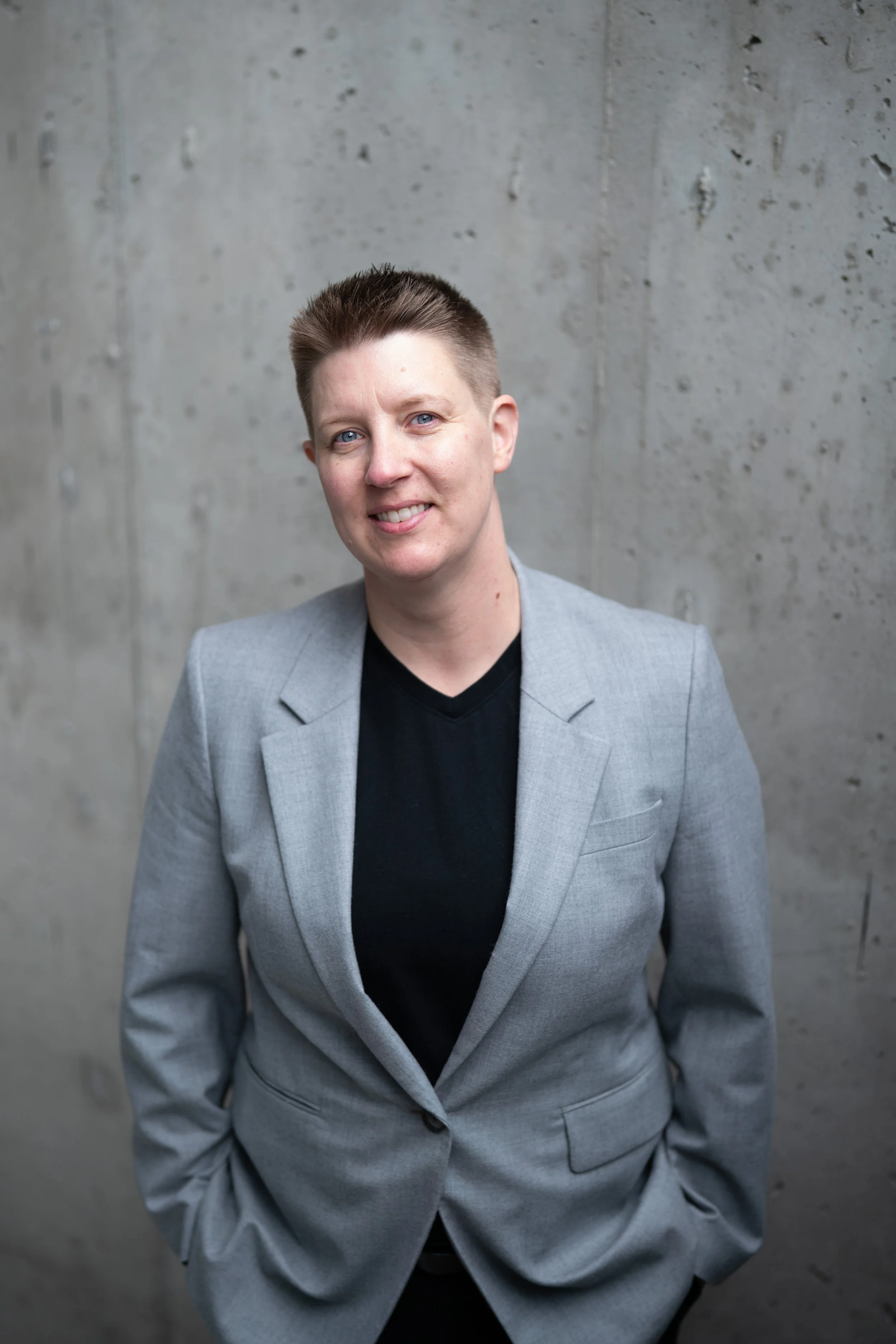 Portrait of a woman with short blonde hair and blue eyes, wearing a gray blazer over a black top, standing against a concrete wall.