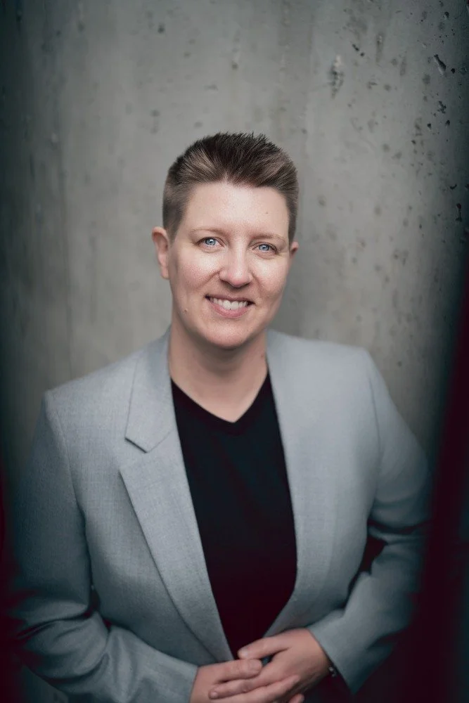 A woman with short brown hair and blue eyes, wearing a light gray blazer over a black top, standing against a concrete wall, smiling at the camera.