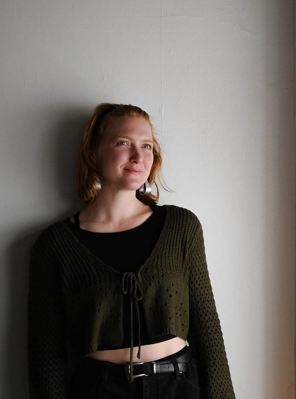 A young woman with shoulder-length red hair standing against a plain white wall, smiling softly and looking slightly to her left.