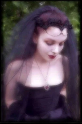 Young woman with dark hair, wearing a black dress and veil, outdoors with greenery.