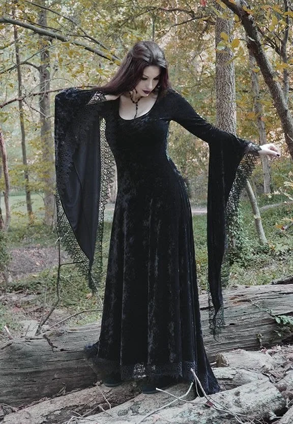 Woman in a long black lace dress with flowing sleeves in a wooded outdoor area.