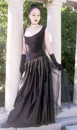 A woman wearing a black dress with a sheer overlay, standing among white columns outdoors.