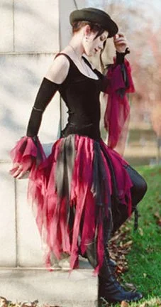 Woman dressed in a black and pink Gothic-inspired outfit with a hat, standing outdoors near a stone wall.