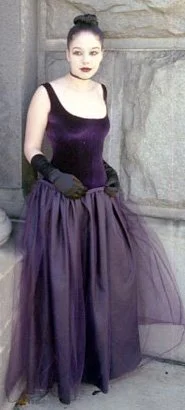 A woman with dark hair styled in an updo, wearing makeup, a purple velvet sleeveless top, purple tulle skirt, and black gloves, standing against a stone wall.