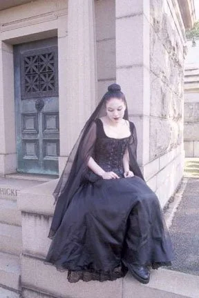 A woman dressed in a black Victorian-style gown with a matching veil sitting on a stone ledge outside a stone building with a decorative door.