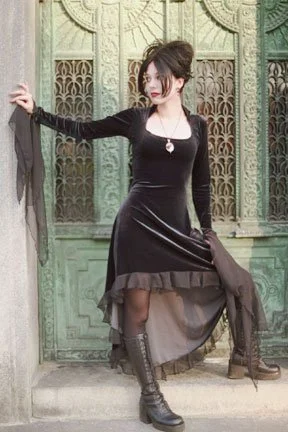 A woman in a black vintage dress with lace-up boots, sitting against an ornate green door, holding a coat.