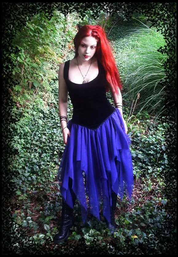 A woman with long red hair wearing a black sleeveless top and a blue layered skirt, standing in a lush green outdoor setting surrounded by plants and foliage.