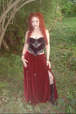 A woman with long red hair standing outdoors among green trees and bushes, wearing a black corset top and a long, flowing red velvet skirt with a slit, paired with black boots.