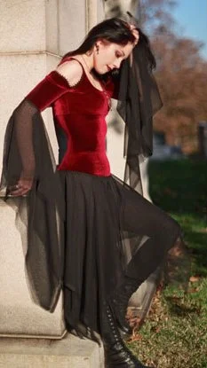 A young woman in gothic attire leaning against a stone structure outdoors, wearing a red velvet top and a long black sheer skirt with black boots.