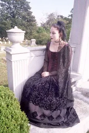Young woman in a black lace dress sitting on a white porch railing with greenery and trees in the background.