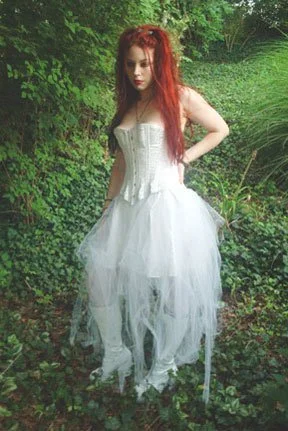 A woman with long red hair wearing a white corset and a flowing white tulle skirt, standing outdoors in a lush green forest or garden.