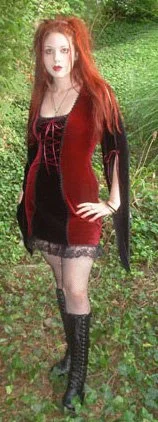 A woman with long red hair standing outdoors on grass, wearing a black and red dress with lace trim and knee-high black lace-up boots.