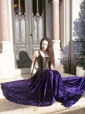 Young woman sitting on steps in front of a building with ornate door, wearing a black top and a long, purple, velvet skirt.