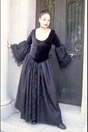 A woman dressed in a black gown with wide, bell sleeves, standing in front of a black metal door.