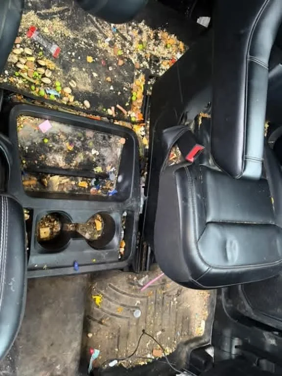 Interior of a car with the center console removed, exposing the gear shifter and scattered trash including candy wrappers and debris on the floor.