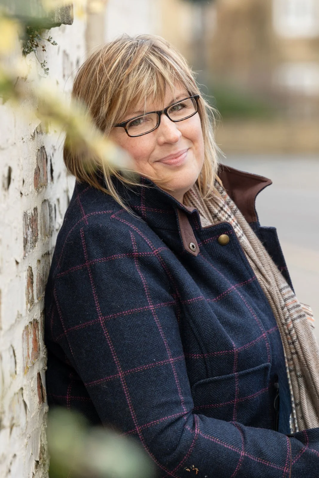 A middle-aged woman with glasses and blonde hair leaning against a brick wall outdoors, smiling softly, wearing a dark plaid coat and a beige scarf.