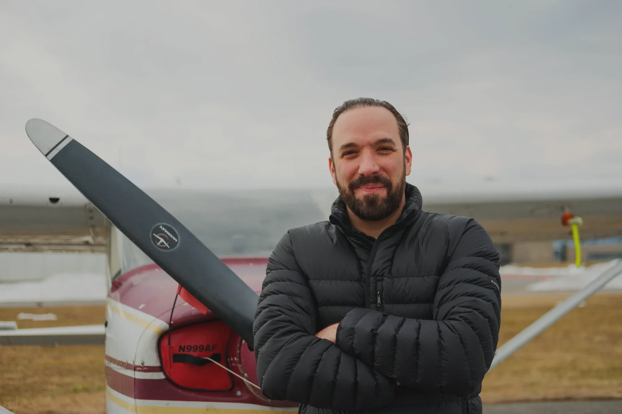 A man with a beard smiling, wearing a black jacket, standing outdoors near a small airplane on a cloudy day.