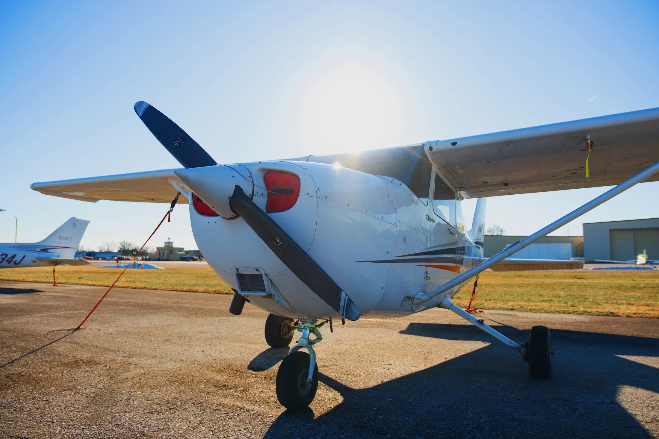 A small propeller airplane parked on the tarmac at an airport with other planes and airport buildings in the background, under a clear blue sky.