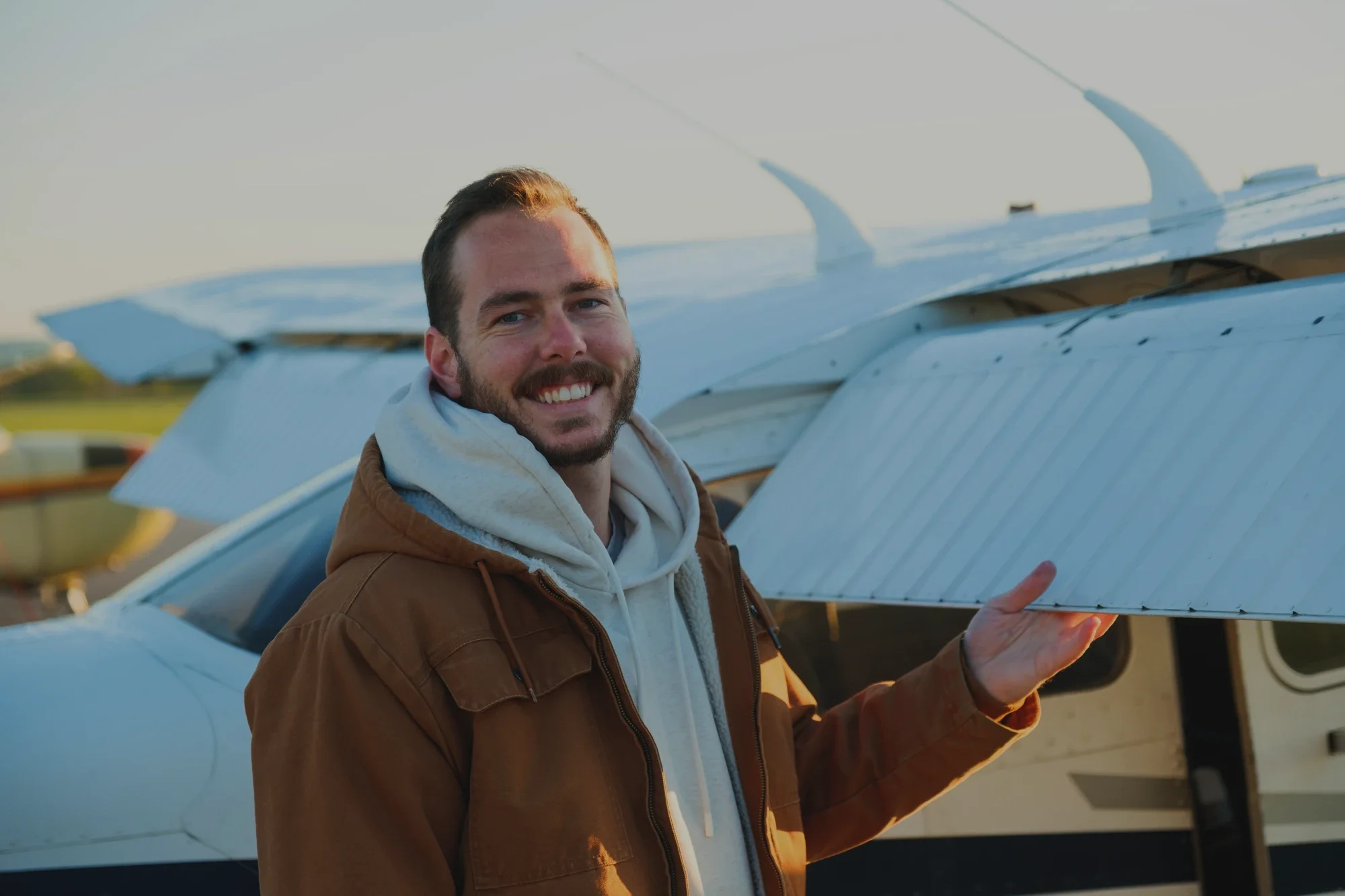 A smiling man with a beard holding a small aircraft model with a white body and wings, standing in front of a real aircraft during sunset.
